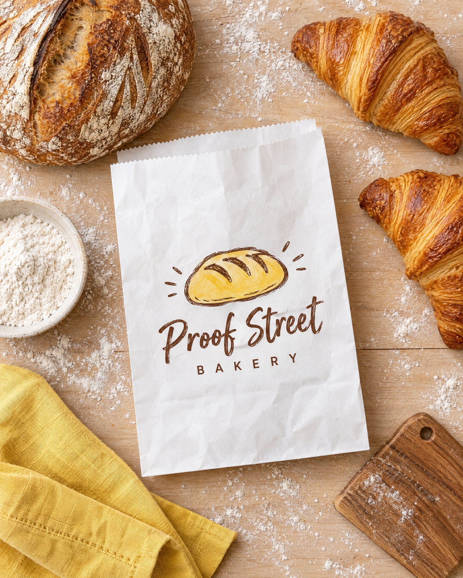 A white paper bag printed with the Proof Street Bakery logo, surrounded by a sourdough loaf, croissants, flour and a yellow napkin, seen from above.