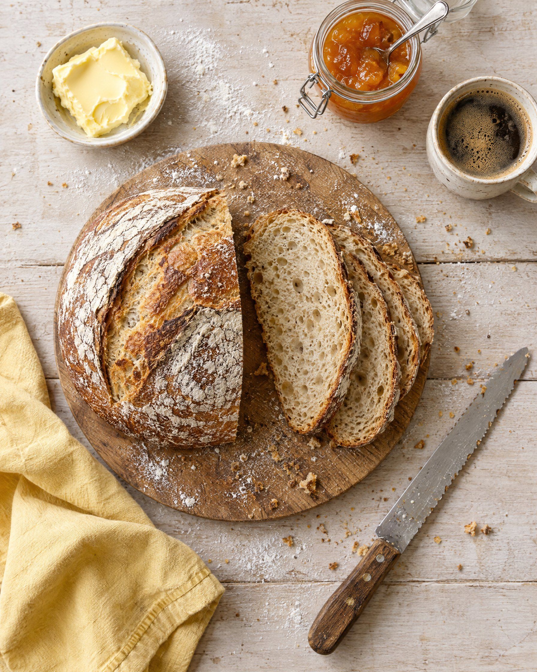 A sliced sourdough loaf on a round board with butter, apricot jam, coffee and a yellow napkin, seen from above.