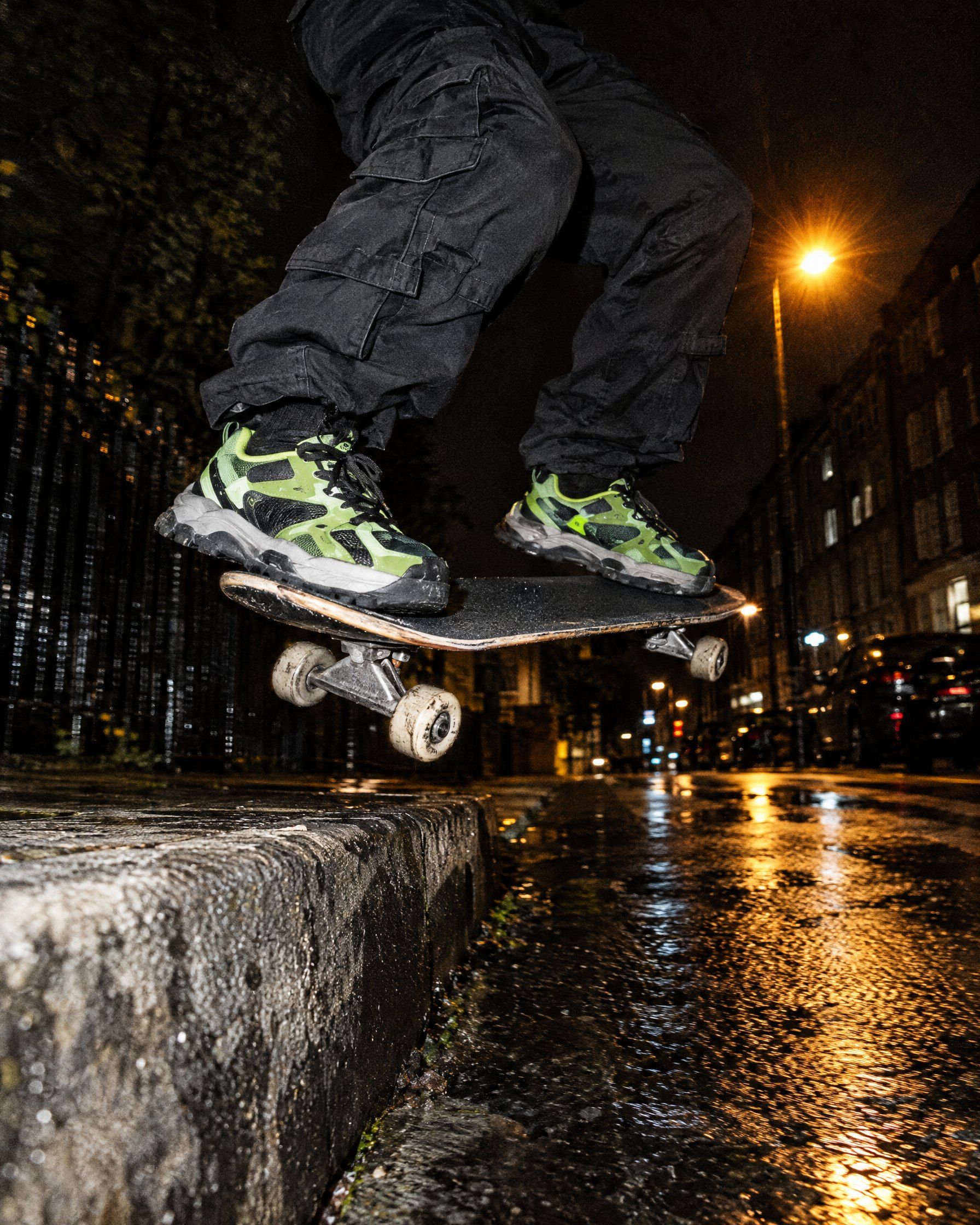 Low angle of a skater in lime trainers and black cargo trousers popping off a kerb at night under a street light.