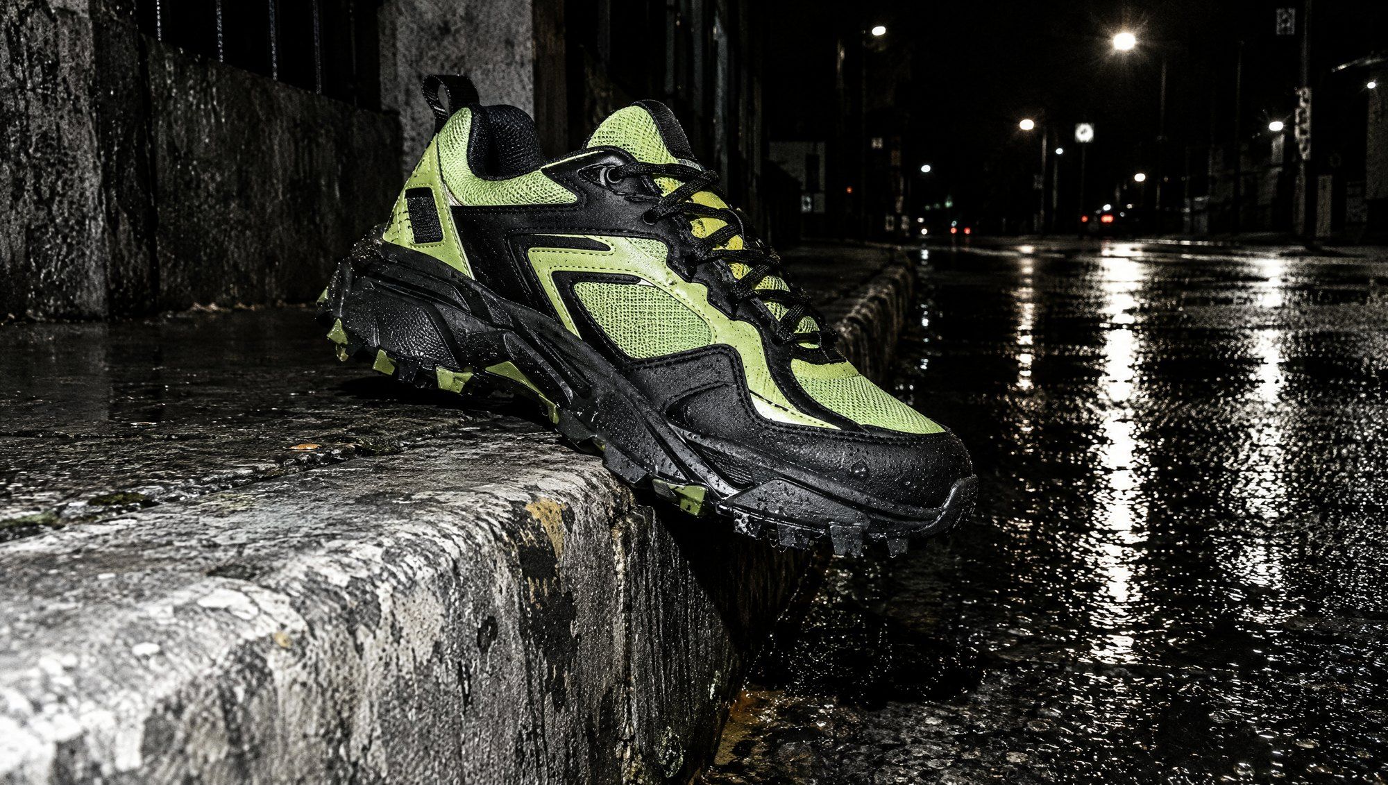 A lime and black trainer balanced on a wet kerb at night, street lights reflecting on the tarmac.