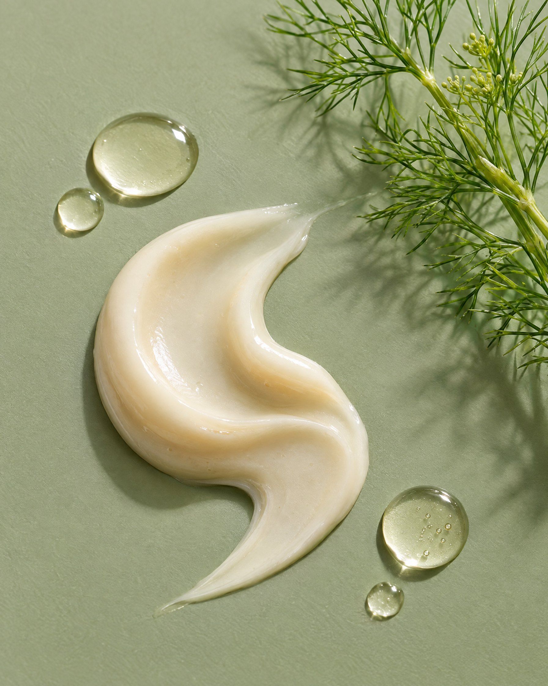 A swirl of cream serum and clear droplets on a sage surface beside a sprig of fennel.